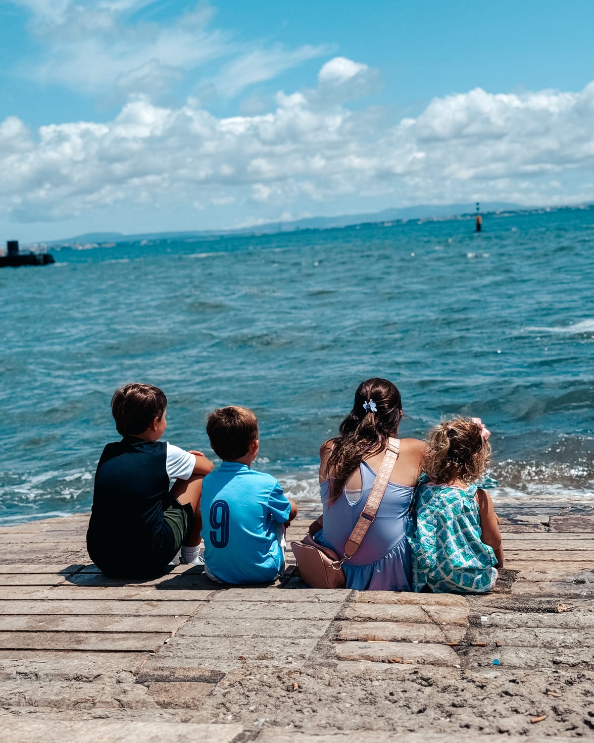A mom and three kids on a stone pier, backs to the camera, looking out over the water in Lisbon